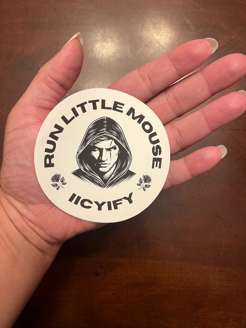 May include: A white round sticker with black text and a black and white illustration of a person wearing a hooded cloak. The text reads "Run Little Mouse Icyify" with a black rose on each side of the text.