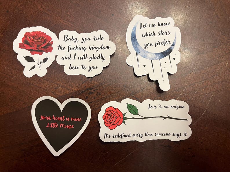 May include: Five stickers with different designs and text. One sticker features a red rose with the text "Baby, you rule the fucking kingdom, and I will gladly bow to you". Another sticker features a black heart with the text "Your heart is mine Little Mouse". A third sticker features a blue crescent moon with stars and the text "Let me know which stars you prefer". A fourth sticker features a red rose with a stem and the text "Love is an enigma. It's redefined every time someone says it". The fifth sticker features a red rose with a stem and the text "Love is an enigma. It's redefined every time someone says it".