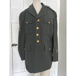 May include: A dark green military jacket with gold buttons and pockets. The jacket has gold stripes on the sleeves and shoulders. The jacket is displayed on a mannequin.