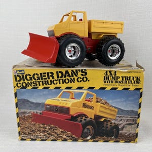 May include: A yellow and red toy dump truck with a dozer blade, sitting on top of its original box. The box features the text "DIGGER DAN'S CONSTRUCTION CO." and "4X4 DUMP TRUCK WITH DOZER BLADE."