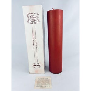 May include: A tall, red pillar candle with a matching box. The box is white with the words "Colonial Pillar Candles" printed in maroon. A small card with text is at the bottom.