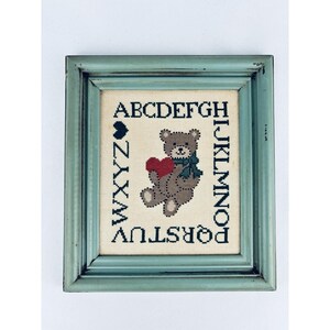 May include: Cross-stitch framed artwork featuring a brown teddy bear holding a red heart, wearing a blue scarf. The alphabet surrounds the bear. The frame is a distressed teal color.