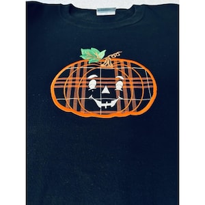 May include: A black crew neck sweatshirt featuring a large embroidered pumpkin graphic on the chest. The pumpkin has an orange and black plaid pattern, a green leaf, and a smiling face with white eyes, a triangular nose, and a stitched mouth.