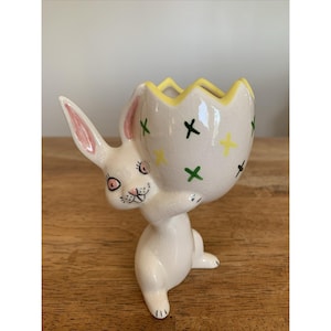 May include: A white ceramic Easter bunny figurine holding a cracked egg. The egg has a yellow rim and is decorated with green and yellow crosses. The bunny has pink-lined ears and a painted face. The figurine is on a wooden surface.