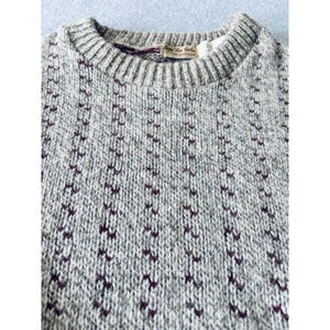 May include: Close-up of a gray knit sweater with a crew neck. The sweater has a textured pattern of small, dark burgundy shapes arranged in vertical rows. The collar features a small tag with text.