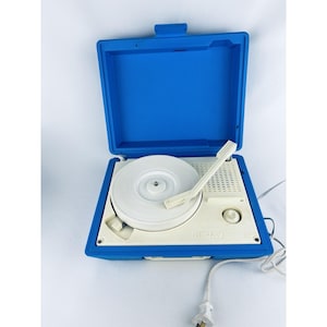 May include: A vintage blue and white portable record player with a white turntable, tone arm, and control knob. The record player is in an open blue case with a handle. A power cord with a plug extends from the unit.