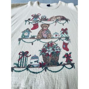 May include: A cream-colored long-sleeve shirt featuring a woven graphic of three shelves decorated with Christmas items including a teddy bear, a cat, wrapped gifts, stockings, a clock, and a basket filled with poinsettia flowers and greenery.