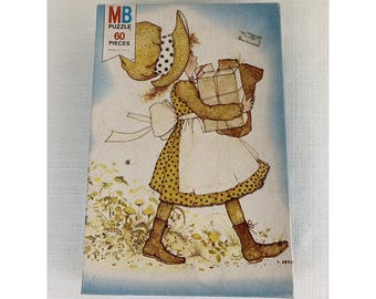 May include: Vintage MB puzzle box featuring a girl in a yellow polka-dot dress and a large sun hat, carrying packages. The box reads "MB PUZZLE 60 PIECES" in blue and white text. The illustration has a soft, nostalgic feel.