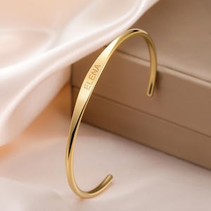以下が含まれることがあります： 前面に「ELENA」と刻印された、金色のカーブしたカフブレスレット。 ブレスレットは、柔らかく明るい色の表面に、ニュートラルなトーンの背景で表示されています。