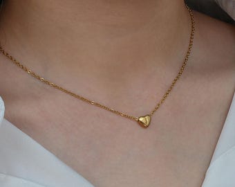 Może przedstawiać: Złoty naszyjnik z delikatnym łańcuszkiem i zawieszką w kształcie serca. Na zawieszce wygrawerowana jest litera "J". Naszyjnik jest noszony na białej koszuli.