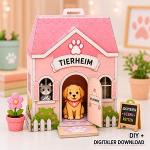 Puede incluir: Un modelo de refugio de animales de papel rosa etiquetado como TIERHEIM con una figura de cachorro dorado en la entrada y un gatito gris en la ventana. La estructura cuenta con una valla blanca, un techo rosa y un letrero que dice ADOPTIEREN LIEBEN RETTEN.