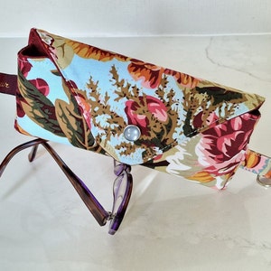 May include: A floral patterned glasses case with a snap closure and a matching strap. The case is open and a pair of purple glasses are inside.