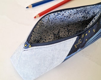May include: A partially open pencil case with a blue zipper, featuring a cityscape design on one side and a light blue fabric on the other. The interior lining has a pattern of white stars on a blue background. Two pencils, one red and one blue, are visible.