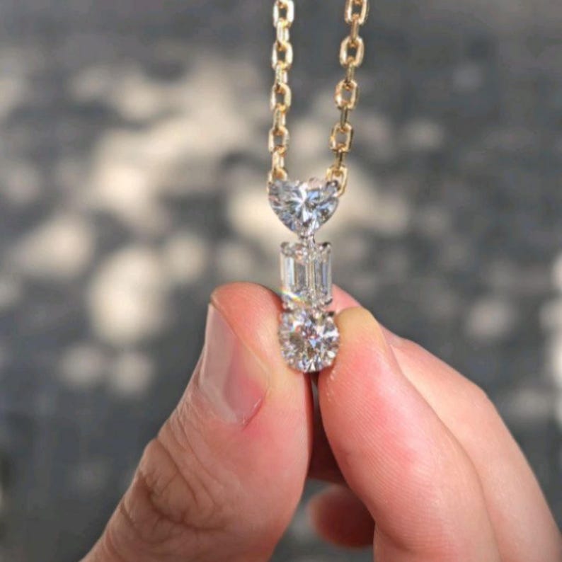 May include: A gold chain necklace with a pendant featuring three clear, sparkling gemstones. The top stone is heart-shaped, the middle is rectangular, and the bottom is round. The necklace is held up by a hand.