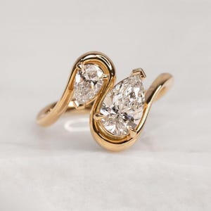 Può includere: Un anello d'oro con due diamanti a forma di pera. Il design dell'anello è a forma libera e fluida, con la fascia d'oro che circonda ogni diamante. L'anello ha una finitura lucida ed è posto su uno sfondo bianco.