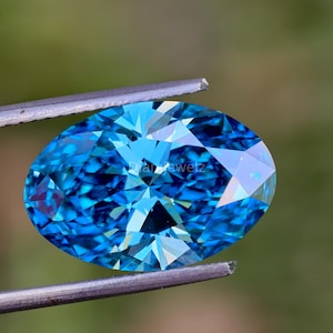 May include: A vibrant blue oval-shaped gemstone with a faceted cut, held by tweezers against a green background.
