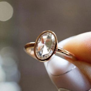 Può includere: Un anello in oro rosa con un diamante taglio ovale. Il diamante è incastonato in una montatura a castone e la fascia è in metallo sottile e lucido. L'anello è mostrato su uno sfondo morbido e sfocato, evidenziando la brillantezza della gemma.
