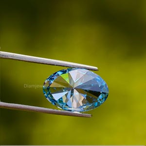 May include: A close-up of a blue oval-shaped gemstone with a faceted cut. The gemstone is held in a pair of tweezers against a green background.