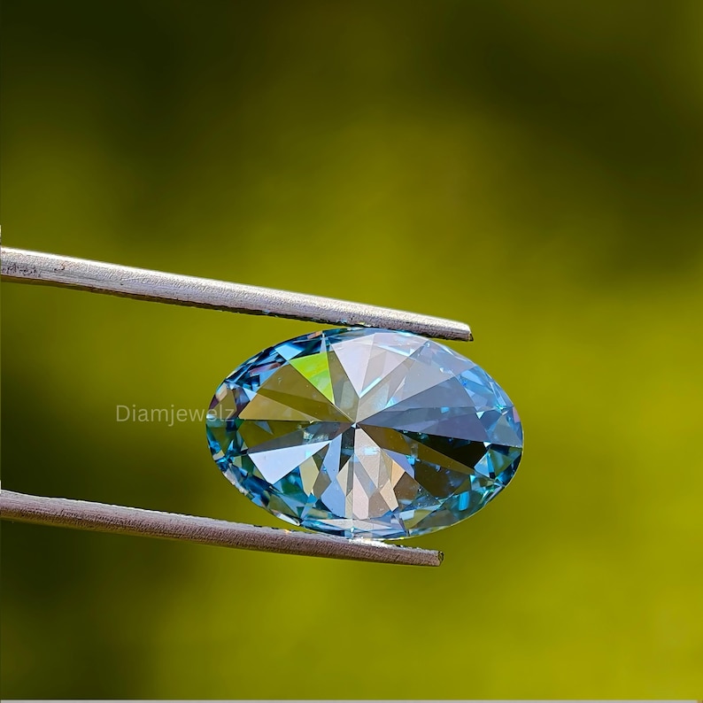 May include: A close-up of a blue oval-shaped gemstone with a faceted cut. The gemstone is held in a pair of tweezers against a green background.