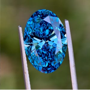 May include: A vibrant blue oval-shaped gemstone with a faceted cut, showcasing a brilliant sparkle. The gemstone is held in a pair of tweezers.