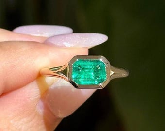 May include: A gold ring featuring a rectangular emerald gemstone. The emerald is set in a bezel setting, and the ring has a delicate band. The emerald is a vibrant green color, and the ring is held up by a hand.