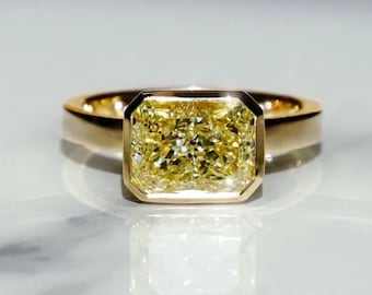 May include: A gold ring featuring a large, rectangular-cut yellow diamond. The diamond is set in a bezel setting, and the band is a simple, polished gold. The ring is displayed on a reflective surface.