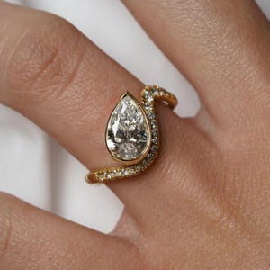 Può includere: Un anello d'oro con un diamante a forma di pera. La fascia è ornata da piccoli diamanti e avvolge il dito. Il design dell'anello è moderno, evidenziando la chiarezza della pietra centrale.