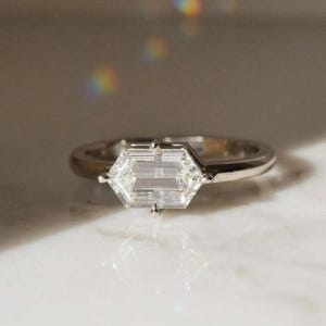 Può includere: Un anello d'argento con una gemma trasparente esagonale. La gemma è il punto focale, incastonata in un design minimalista. L'anello è su una superficie bianca, con un'illuminazione soffusa e un sottile effetto arcobaleno.