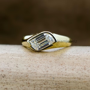 Può includere: Un anello d'oro con una gemma rettangolare e trasparente. La gemma è incastonata in una lunetta, e la fascia dell'anello è liscia e lucida. L'anello è esposto su una superficie marrone chiaro. Il testo "Diamjewelz" è visibile.
