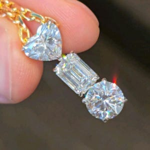 May include: A gold chain necklace with a pendant featuring three clear, sparkling diamonds. The diamonds are heart-shaped, emerald-cut, and round. The necklace is held by a person's fingers.