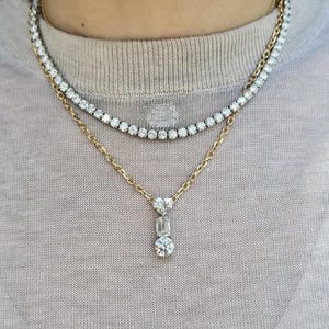 May include: A layered necklace set featuring a tennis necklace and a gold chain with a pendant. The pendant has a heart-shaped stone, an emerald-cut stone, and a round stone. The jewelry is set against a neutral background.