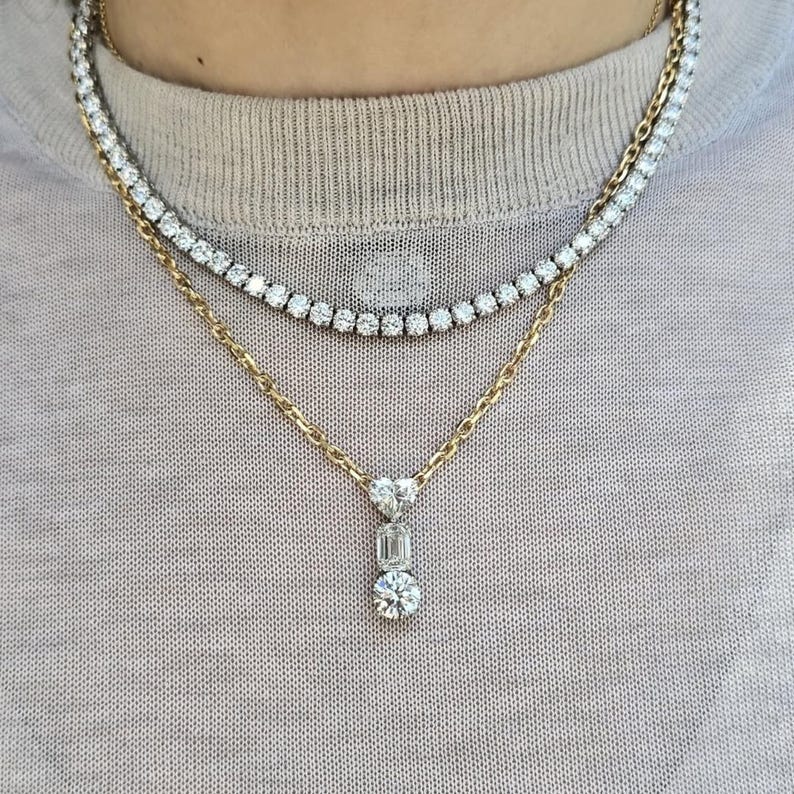 May include: A layered necklace set featuring a tennis necklace and a gold chain with a pendant. The pendant has a heart-shaped stone, an emerald-cut stone, and a round stone. The jewelry is set against a neutral background.