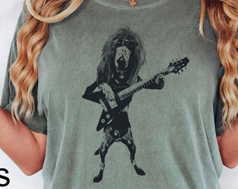 May include: A moss green short-sleeve t-shirt featuring a black graphic print of a dog with long hair playing an electric bass guitar. The shirt has a crew neckline and a relaxed fit, with the word Moss printed in black text on the lower left side.