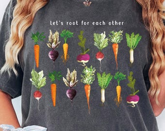 May include: A charcoal gray short-sleeve t-shirt featuring a graphic print of various colorful root vegetables including carrots, beets, and radishes. The text "Let's root for each other" is printed in white above the vegetable illustration.