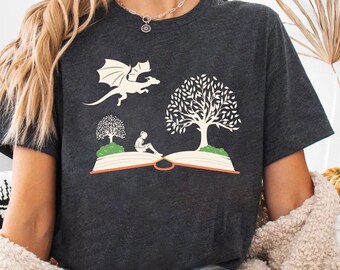 May include: Dark gray t-shirt featuring a whimsical design with a dragon, trees, and a person reading on an open book. The graphic is in a cream color. The shirt is a comfortable, casual wear.