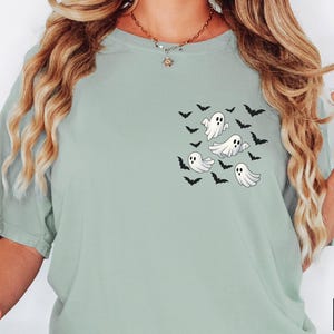 May include: A light green t-shirt featuring a pocket design with cartoon ghosts and bats. The shirt is a solid color and has a relaxed fit. The design is in black and white.