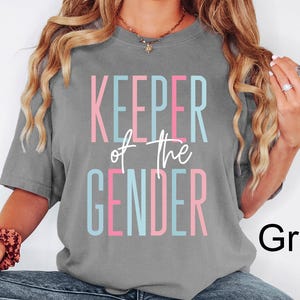 May include: A grey t-shirt with the text "KEEPER of the GENDER" in pink, blue, and white. The shirt is a crew neck style.