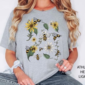 May include: Light grey t-shirt featuring a floral and bee design. The design includes yellow sunflowers, white daisies, green leaves, and black and yellow bees. The text "ATHLETHICH HEATHER LIGHT GREY" is printed on the right side of the image.