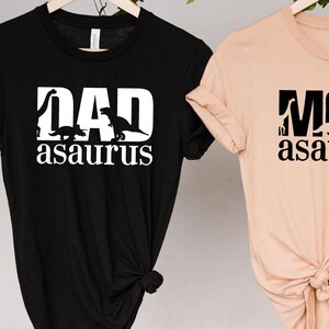 May include: Two black and beige t-shirts with a dinosaur graphic and the text "DAD asaurus" and "MOM asaurus".