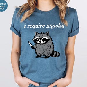 May include: Heather deep teal t-shirt with the text "i require snacks" above a cartoon raccoon holding a knife. The raccoon is gray and black with a white face and a striped tail. The shirt is a solid color.