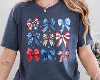 May include: Dark blue t-shirt featuring a grid of watercolor bows in red, white, and blue. The bows have star and stripe patterns, perfect for patriotic celebrations. The shirt is made of a soft material.