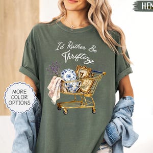 May include: Olive green t-shirt with the text "I'd Rather Be Thrifting." The graphic features a gold shopping cart filled with vintage items, including a blue and white plate, a framed picture, and lavender. The shirt is made of hemp.