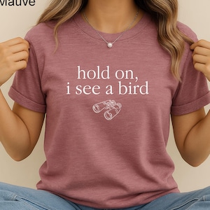 May include: Mauve-colored t-shirt with the text "hold on, i see a bird" and a graphic of binoculars. The shirt is short-sleeved and made of a soft material. The text and graphic are in white.
