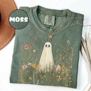 May include: A moss green t-shirt featuring a ghost in a field of wildflowers. The ghost is white with black eyes and a simple design. The shirt has a crew neck and short sleeves. A silver necklace with a pendant is visible.