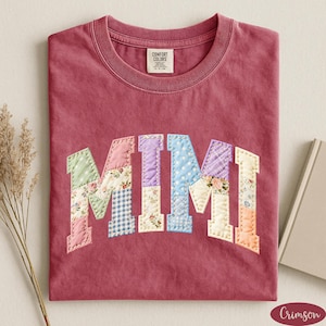 May include: A crimson-colored t-shirt with the word "MIMI" appliqued across the front. The letters are made of patchwork fabric in various floral and patterned designs. The shirt has a crew neck and a tag that reads "Comfort Colors".