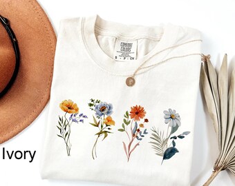 May include: An ivory t-shirt featuring a graphic print of four distinct wildflower bouquets in shades of orange, blue, and green. The shirt has a crew neckline and a Comfort Colors brand label at the inner neck. A gold-toned necklace rests on top.