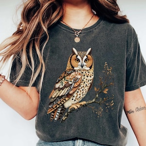 May include: Dark gray t-shirt featuring a detailed illustration of an owl perched on a branch. The owl has brown, tan, and cream-colored feathers. The shirt is a crew neck style.