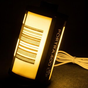 May include: A film canister lamp emitting a warm yellow light. The black canister features the text "PORTRA 400 EXP 36" and a barcode design. A white cable extends from the lamp.