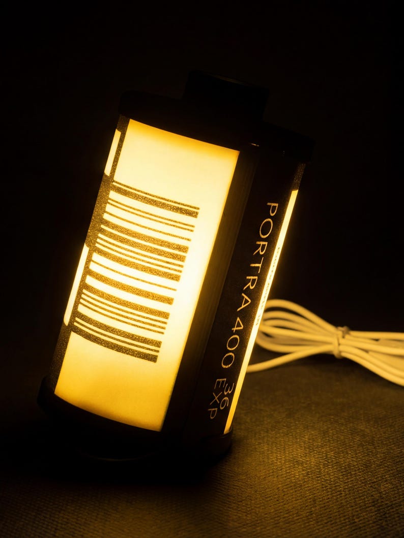 May include: A film canister lamp emitting a warm yellow light. The black canister features the text "PORTRA 400 EXP 36" and a barcode design. A white cable extends from the lamp.