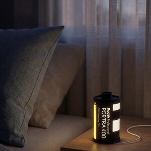 May include: A black and yellow Kodak Professional Portra 400 film canister lamp on a wooden bedside table. The lamp is illuminated, displaying the text "Kodak Professional Portra 400 Color Negative Film". A white cable extends from the lamp.